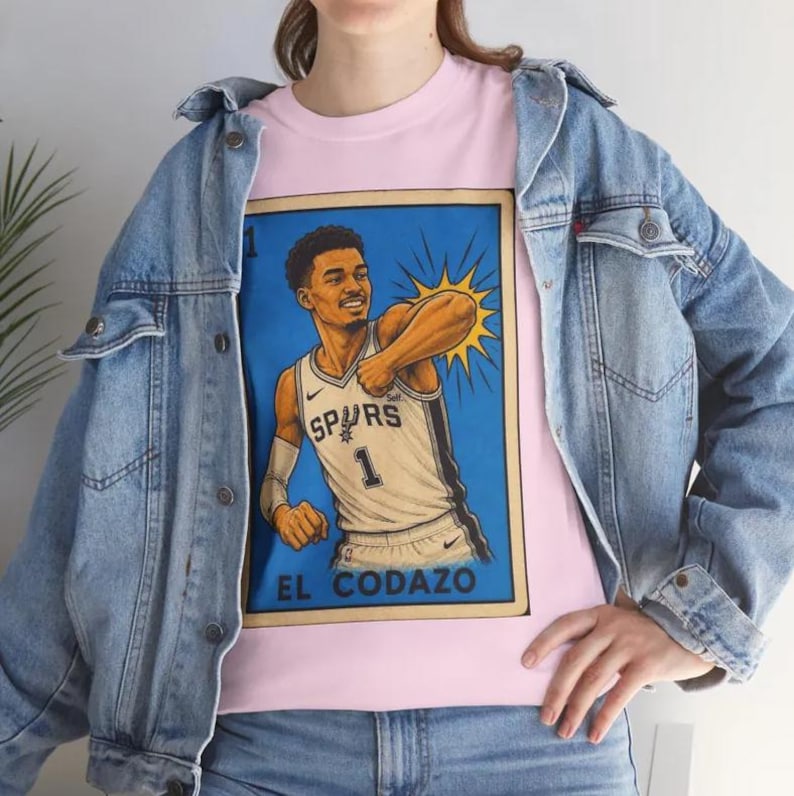 May include: A pink t-shirt with a graphic of a basketball player in a Spurs jersey with the number 1. The graphic is in a lottery ticket style, with the text "EL CODAZO" at the bottom. The person is wearing a light blue denim jacket.