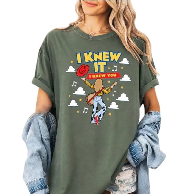 Pu&ograve; includere: Una t-shirt verde oliva con la grafica di una persona dai capelli lunghi che suona la chitarra, indossando una camicia gialla e pantaloni con stampa muccata. Il testo recita I KNEW IT e I KNEW YOU, circondato da un cappello rosso, nuvole, stelle e note musicali.