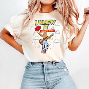 Pu&ograve; includere: Una t-shirt a maniche corte color crema con la grafica di una persona che suona la chitarra, indossando una camicia gialla e pantaloni con stampa muccata. Il testo recita I KNEW IT e I KNEW YOU, circondato da un cappello rosso e nuvole.