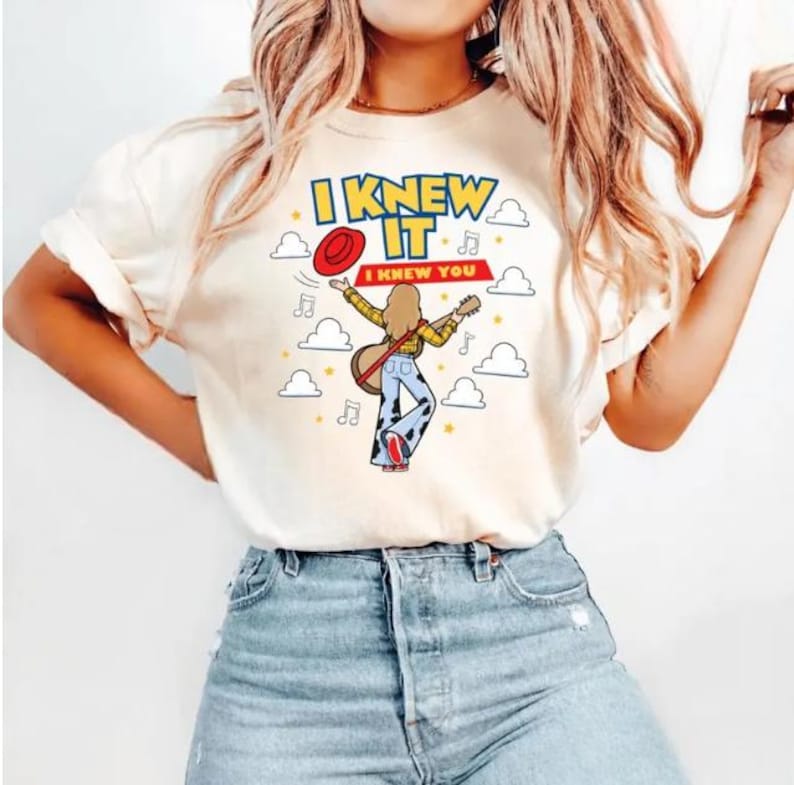 Pu&ograve; includere: Una t-shirt a maniche corte color crema con la grafica di una persona che suona la chitarra, indossando una camicia gialla e pantaloni con stampa muccata. Il testo recita I KNEW IT e I KNEW YOU, circondato da un cappello rosso e nuvole.