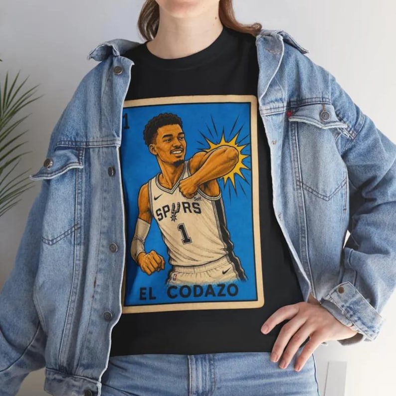 May include: Black t-shirt with a basketball player in a Spurs jersey, number 1, set against a blue background. The text "EL CODAZO" is visible. The shirt is worn with a denim jacket.
