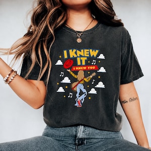 Pu&ograve; includere: Una t-shirt a maniche corte grigio antracite con una stampa grafica di una persona con i capelli lunghi che suona la chitarra. Il testo recita I KNEW IT e I KNEW YOU. Il design include un cappello rosso, nuvole bianche, stelle gialle e note musicali.