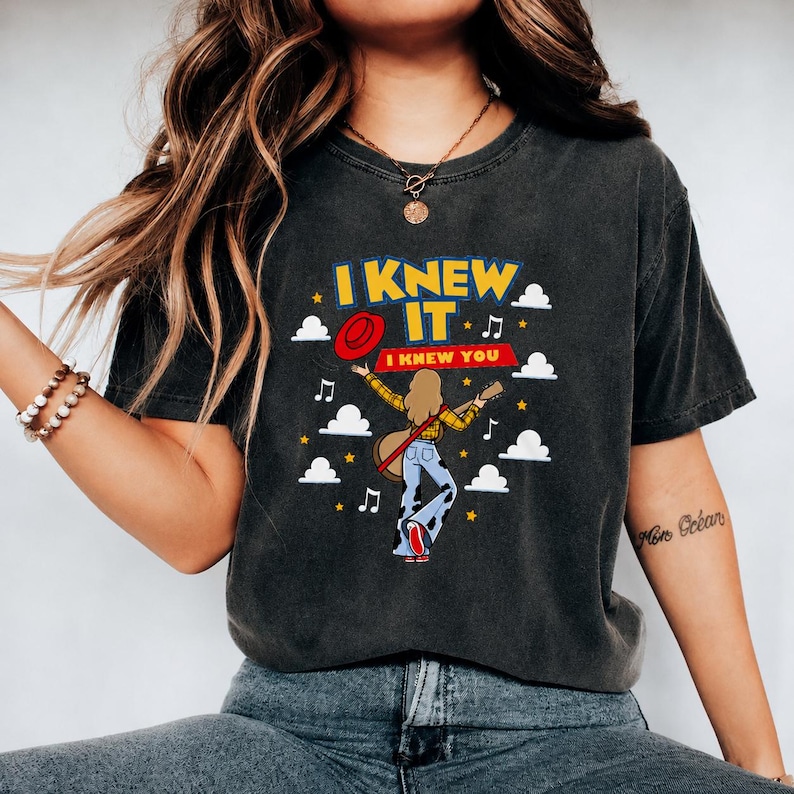 Pu&ograve; includere: Una t-shirt a maniche corte grigio antracite con una stampa grafica di una persona con i capelli lunghi che suona la chitarra. Il testo recita I KNEW IT e I KNEW YOU. Il design include un cappello rosso, nuvole bianche, stelle gialle e note musicali.