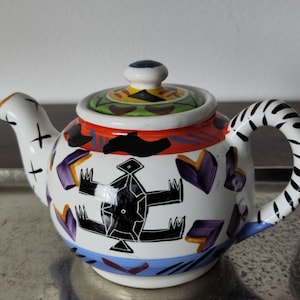 Op de afbeelding: Decoratieve keramische theepot met een witte basis, met een zwart, rood en paars geometrisch ontwerp. De handgreep heeft een zwart-wit zebrapatroon. Het deksel is bekroond met een kleine blauwe knop.