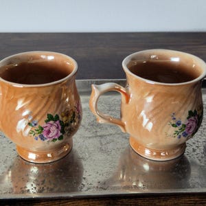 Pode incluir: Duas canecas de cerâmica pêssego vintage com desenhos florais, colocadas numa bandeja prateada. Cada caneca tem uma pega e é decorada com flores rosa, roxas e azuis. As canecas são exibidas numa superfície refletora.
