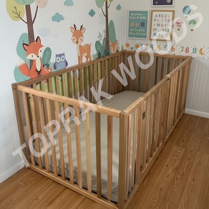 以下が含まれることがあります： 垂直なスラットとヒンジ付きゲートを備えた、明るい色の木製ベビーサークル。ベビーサークルにはマットレスが入っており、広葉樹の床に置かれています。背景には、森の動物と教育ポスターの壁画があります。