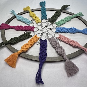 May include: A collection of colorful macrame keychains arranged in a circular pattern. Each keychain features a woven design in various colors, including pink, blue, yellow, and green, with a tassel at the end and a silver keyring.