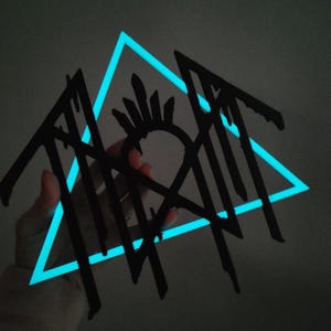 May include: A black, jagged, stylised logo featuring sharp, dripping lines and a central circular emblem with radiating spikes, layered over a glowing, bright cyan triangular frame that illuminates the dark background behind the design.
