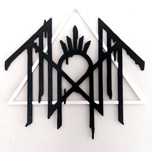May include: A wall-mounted decorative piece featuring a white triangular frame layered behind a black, stylized, dripping-effect emblem. The black design includes a central arched structure topped with spikes and flanked by jagged, downward-dripping lines.