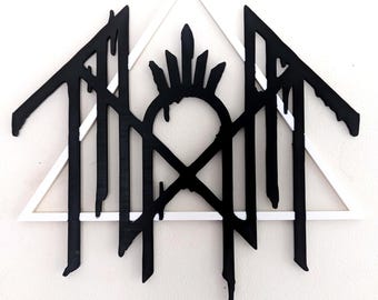 Puede incluir: Una pieza decorativa de pared que presenta un marco triangular blanco superpuesto a un emblema negro con efecto de goteo. El diseño negro incluye una estructura central arqueada con puntas y líneas irregulares que caen hacia abajo.
