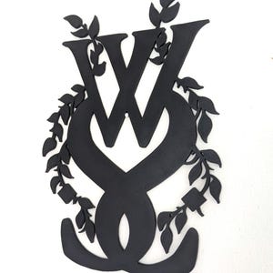 May include: A matte black wall decoration featuring the letter W intertwined with a stylized heart shape, surrounded by a decorative border of leafy vines. The design is flat and constructed from a solid material with a textured surface finish.