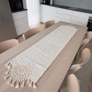 Può includere: Runner da tavolo rettangolare color crema con un design circolare all'uncinetto e nappe. Il runner è posto su un tavolo di legno chiaro, circondato da sedie di legno. L'ambientazione suggerisce una sala da pranzo o una cucina.