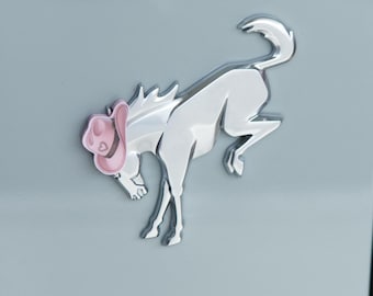 Peut inclure: Un emblème de cheval chromé avec un chapeau de cow-boy rose. L'emblème est fixé sur une surface gris clair. Le cheval se cabre sur ses pattes arrières. Le texte "DRESS UP MY BRONCO" est en bas.