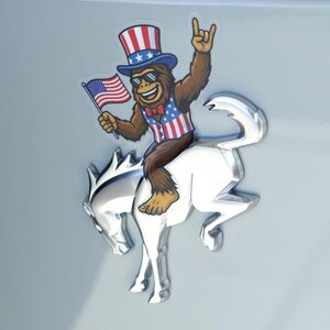 May include: A sticker featuring a cartoon Bigfoot wearing sunglasses, a patriotic vest, and a top hat, holding an American flag and giving a rock-on hand gesture. The Bigfoot is riding a silver horse emblem.
