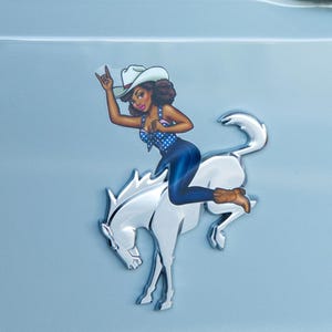 May include: A chrome-colored emblem featuring a woman in a cowboy hat and attire riding a white horse. The woman is wearing a blue polka-dot top, blue jeans, and brown boots. The emblem is mounted on a light blue surface.