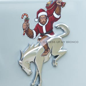 May include: A decal featuring a brown Bigfoot character wearing a red and white Santa suit, holding a candy cane and a red sack of gifts, positioned over a silver metallic bucking horse emblem with the text DRESS UP MY BRONCO printed nearby.