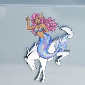 May include: A sticker featuring a mermaid with long pink hair, a seashell top, and an iridescent blue tail, positioned over a chrome-colored horse emblem. The text DRESS UP MY BRONCO is printed in white at the bottom of the image.