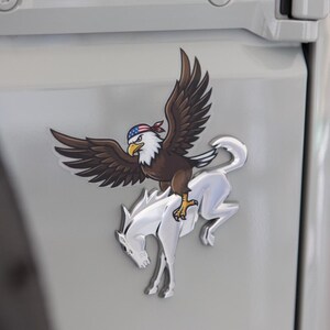 May include: A metal emblem featuring a brown eagle with a red, white, and blue bandana riding a silver horse. The emblem is affixed to a light gray surface. The text "© DRESS UP MY BRONCO" is at the bottom.