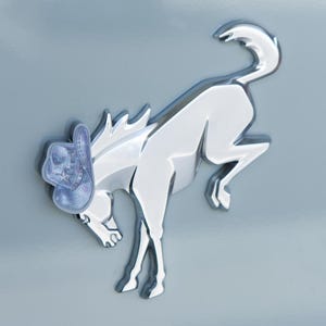 May include: A chrome-colored emblem of a rearing horse wearing a light blue cowboy hat with a star design. The emblem is mounted on a light blue surface. The text "DRESS UP MY BRONCO" is at the bottom.