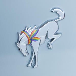 May include: A chrome-finished, metallic horse emblem in a bucking pose, featuring a vibrant rainbow-colored bandana tied around its neck. The emblem is mounted on a light blue surface and includes the text DRESS UP MY BRONCO at the bottom.