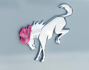 Peut inclure: Un emblème chromé d'un cheval cabré avec un chapeau de cowboy rose. Le chapeau a une étoile et une bordure argentée. L'emblème est sur une surface gris clair. Le texte "DRESS UP MY BRONCO" est en bas.
