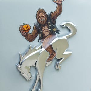 May include: A clear sticker featuring a brown, furry Bigfoot creature wearing a black vest, holding an orange jack-o'-lantern, and riding a silver, metallic, bucking horse emblem. The text DRESS UP MY BRONCO is printed at the bottom.