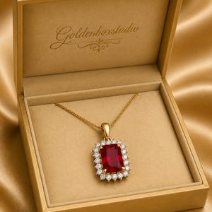 Könnte beinhalten: Eine goldene Halskette mit einem rechteckigen, roten Edelsteinanhänger, umgeben von kleinen, klaren Steinen, präsentiert in einer goldenen Schatulle. Die Schatulle ist geöffnet und zeigt die Halskette vor einem beigen Innenraum. Die Schatulle liegt auf goldenem Satin.