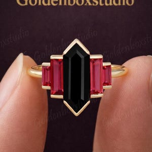 May include: A gold ring with a central black gemstone and rectangular red gemstones. The ring is held up by a person's fingers. The text "Goldenboxstudio" is in the background. The ring's design is geometric.