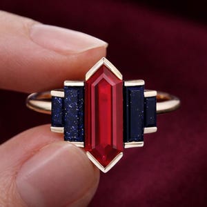 May include: A gold ring with a central, rectangular red gemstone and two dark blue rectangular side stones. The ring has a geometric design and a gold band. The text "Goldenboxstudio" is at the bottom.
