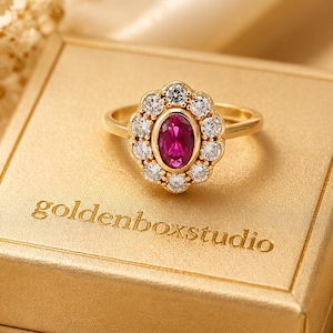 May include: A gold ring featuring a central oval-cut deep pink gemstone surrounded by a halo of small round clear stones. The ring rests on a square gold box with the text goldenboxstudio printed in a serif font on the top surface.