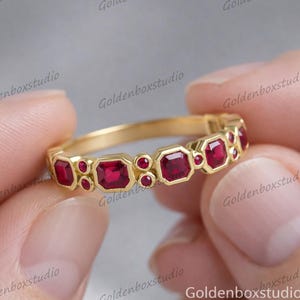 Puede incluir: Un anillo de oro con piedras rojas cuadradas y redondas alternas. El anillo tiene una banda delicada y se sostiene entre dos dedos.