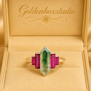 May include: A gold ring featuring a central green and white gemstone, with rectangular pink gemstones on either side. The ring is presented in a gold box, with the text "Goldenboxstudio". The box rests on a gold surface.