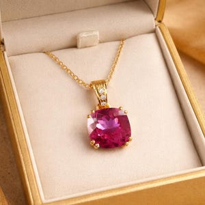Puede incluir: Un collar de oro con un gran colgante cuadrado de piedra preciosa rosa. El colgante está engastado en un marco dorado y rodeado de piedras preciosas más pequeñas y transparentes. El collar se muestra en una caja de joyería dorada.