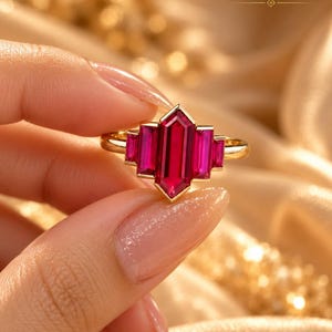 May include: A gold ring featuring a central elongated hexagonal red gemstone flanked by two smaller baguette-cut red gemstones on each side, all set in a polished gold band. The text GoldenBoxStudio is visible in the upper right corner.