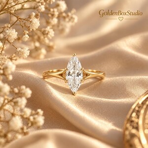 May include: A gold ring featuring a marquise-cut clear gemstone set in a prong setting with a split shank design. The text GoldenBoxStudio with a heart icon is visible in the upper right corner. The ring is positioned on a soft, draped fabric.