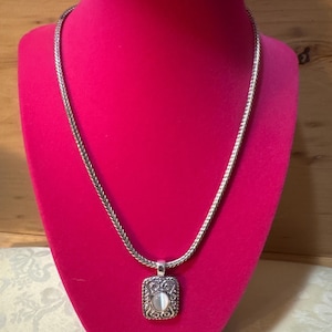 May include: A silver-toned necklace featuring a thick, woven chain and a rectangular pendant. The pendant has an ornate, textured border with a central, smooth, oval-shaped white stone. The necklace is displayed on a bright pink velvet bust.