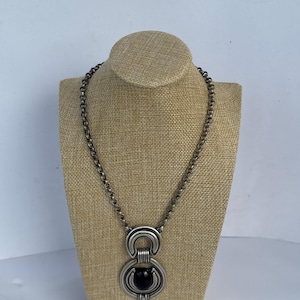 Peut inclure: Un collier en métal argenté composé d'une chaîne épaisse et d'un grand pendentif circulaire. Le pendentif se compose de deux arches semi-circulaires superposées reliées par des barres verticales, avec une pierre noire ovale lisse au centre.