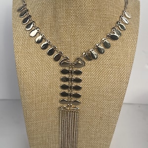 May include: A gold-tone necklace featuring a series of small, flat, oval-shaped metal discs along the chain. The centre of the necklace displays a vertical arrangement of six stacked oval discs, terminating in a long, dangling fringe of thin gold chains.