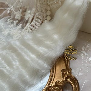 May include: A long, flowing, white faux fur tail, with a soft, textured appearance. The tail is positioned next to a gold-coloured, ornate mirror frame. The words "soul factory" and "White" are visible on the frame.