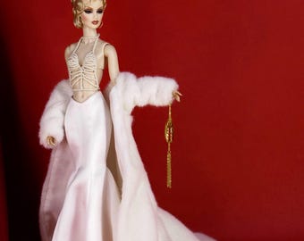 Könnte beinhalten: Eine Sammlerpuppe mit welligem blondem Haar, bekleidet mit einem weißen perlenbesetzten Neckholder-Top und einem bodenlangen weißen Satinrock. Die Puppe trägt einen langen weißen Kunstpelzmantel und hält eine kleine goldene Kettentasche.