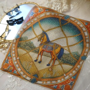 May include: A square silk scarf featuring a central illustration of a brown horse standing within a golden chain-link frame against a starry blue sky background, accompanied by a gold-toned handheld mirror with a reflective surface.
