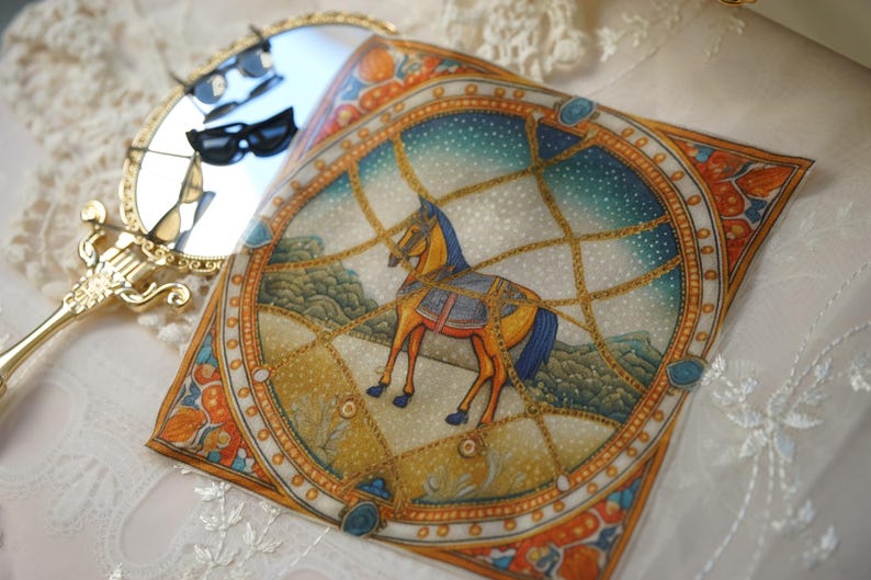 May include: A square silk scarf featuring a central illustration of a brown horse standing within a golden chain-link frame against a starry blue sky background, accompanied by a gold-toned handheld mirror with a reflective surface.