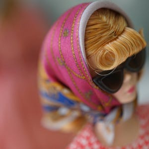 May include: A doll with blonde hair styled in a side-swept fringe, wearing black cat-eye sunglasses and a vibrant pink headscarf featuring gold chain patterns and blue accents. The doll is dressed in a red and white checkered top with a light gray trim.