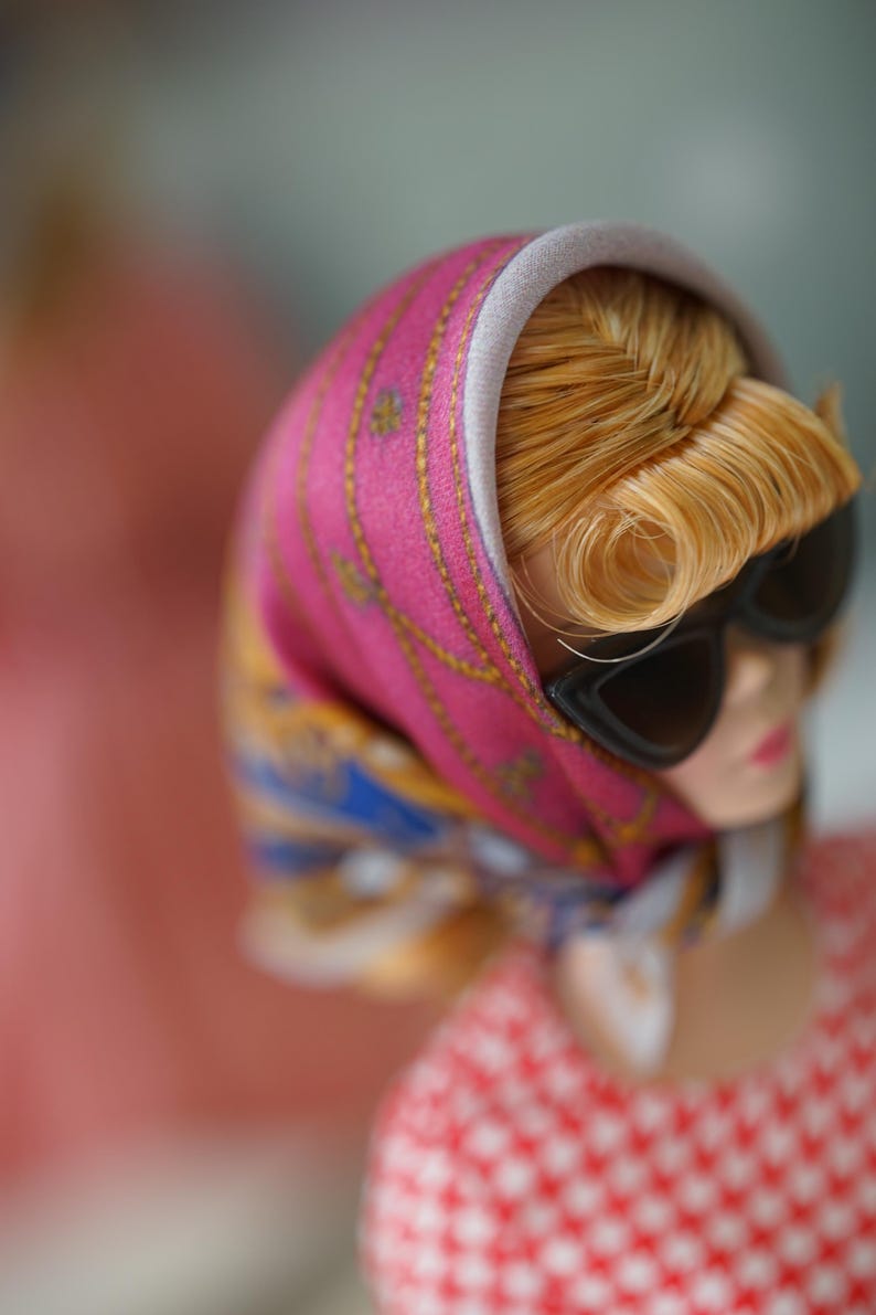 May include: A doll with blonde hair styled in a side-swept fringe, wearing black cat-eye sunglasses and a vibrant pink headscarf featuring gold chain patterns and blue accents. The doll is dressed in a red and white checkered top with a light gray trim.