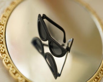Könnte beinhalten: Eine schwarze Sonnenbrille mit abgerundetem Rahmendesign, platziert auf einem runden Spiegel mit einem dekorativen goldfarbenen Metallrand. Die Sonnenbrille spiegelt sich in der Spiegelfläche und zeigt die Vorder- und Seitenansicht der Fassung.