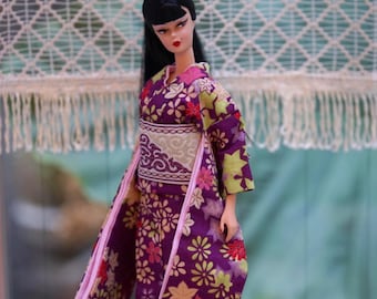 Könnte beinhalten: Eine Puppe mit schwarzem, hochgestecktem Haar, bekleidet mit einem violetten Kimono mit mehrfarbigem Blumen- und Blattmuster. Das Kleidungsstück umfasst einen breiten, strukturierten cremefarbenen Obi-Gürtel mit dunkelbrauner Bordüre.
