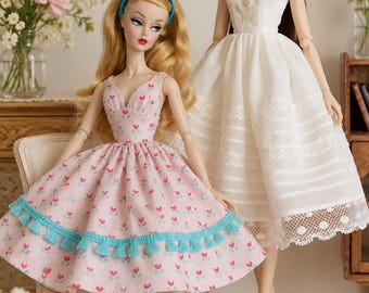 Könnte beinhalten: Zwei Modepuppen stehen zusammen. Eine Puppe hat blondes Haar, ein blaues Stirnband und ein rosa Kleid mit kleinem Herzmuster und blauer Quastenborte, kombiniert mit blauen Absätzen. Die andere Puppe hat langes braunes Haar und ein weißes Kleid.