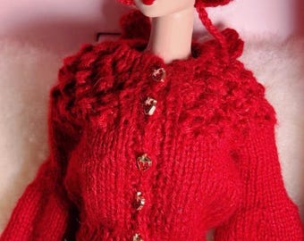 Könnte beinhalten: Eine Puppe trägt eine leuchtend rote, handgestrickte Strickjacke mit fünf kleinen goldfarbenen erdbeerförmigen Knöpfen und eine passende rote Häkelmütze. Die Puppe hat schwarzes, lockiges Haar und trägt einen flauschigen hellrosa Rock mit bunten Sprenkeln.