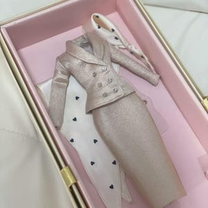 以下が含まれることがあります： シャンパンカラーのきらめくダブルブレストのブレザーと、お揃いのペンシルスカートがセットになったミニチュアサイズのドール用スーツ。小さな黒いハート柄があしらわれた白いスカーフまたはアクセサリーが付属しています。