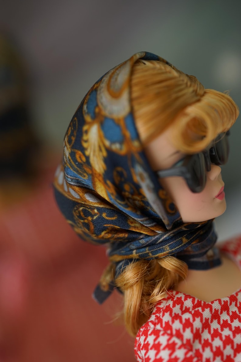 May include: A doll with blonde hair styled in a side-swept fringe, wearing dark-tinted sunglasses and a navy blue headscarf featuring a gold and white ornate pattern. The doll is dressed in a red and white houndstooth patterned garment.